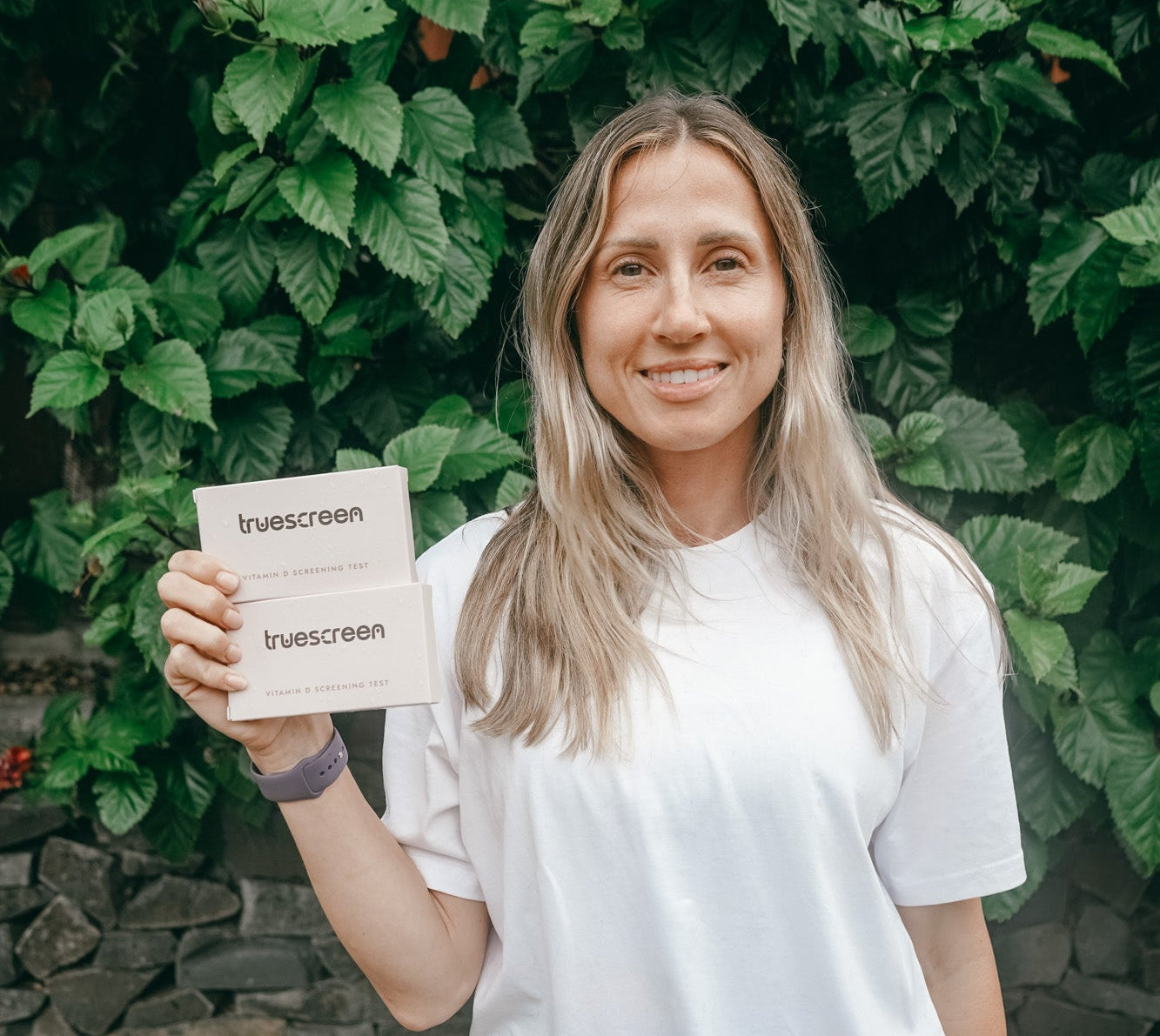 Woman holding a Vitamin D screening test with 'truescreen' branding in front of green foliage.

