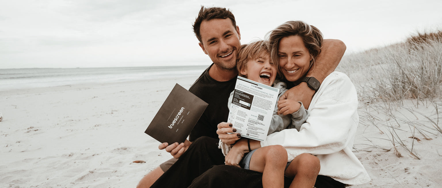 Family of three sitting on a sandy beach holding a TrueScreen FIT Bowel Cancer Screening Test